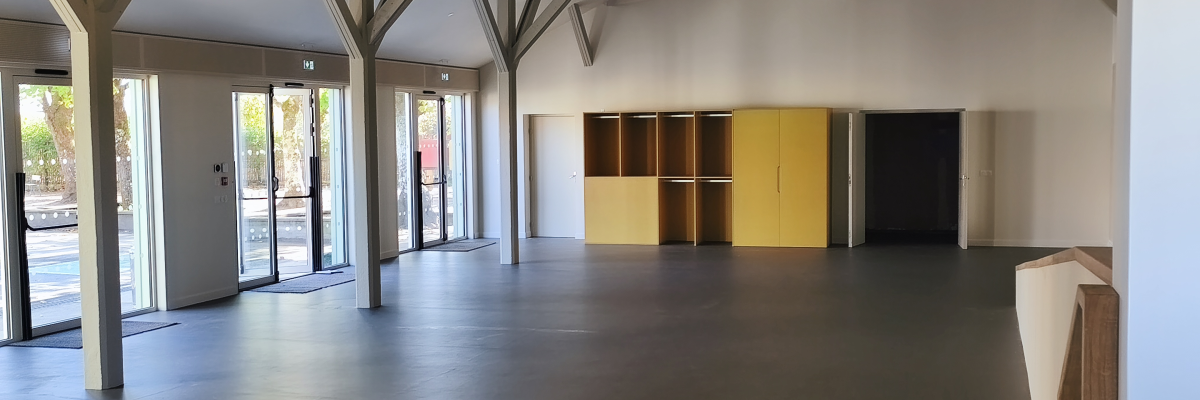 Salle spacieuse avec plafond haut, piliers en bois, bancs, porte, et grande baie vitrée.