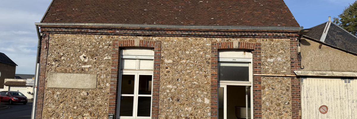 Bâtiment en pierre avec toit en tuiles, porte blanche ouverte, fenêtre à droite, porte de garage blanche à gauche.