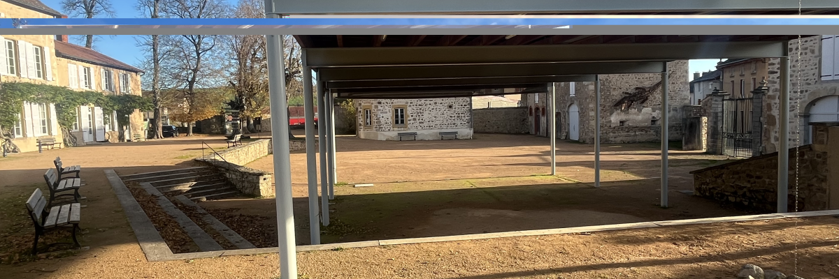 Structure avec toit en appentis, banc, sol terreux, bâtiments en arrière-plan.