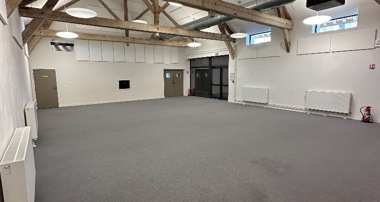 Salle vide avec piliers, sol gris, lumières suspendues et fenêtres en haut.
