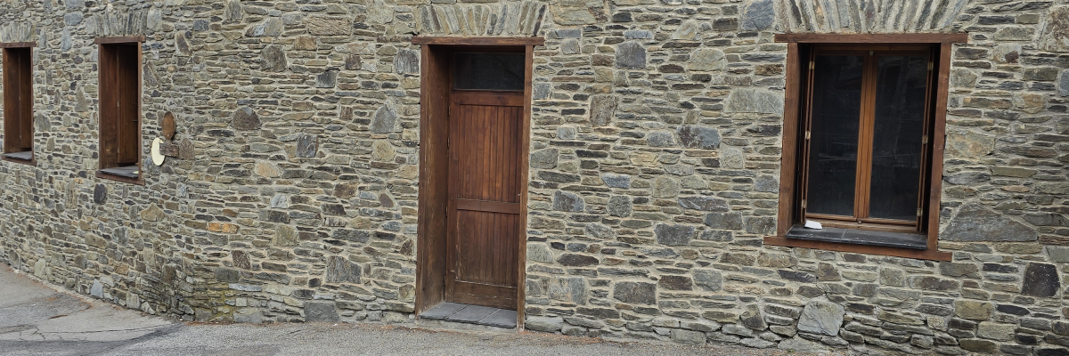 Maison en pierre avec trois fenêtres et une porte en bois, cour asphaltée fissurée.
