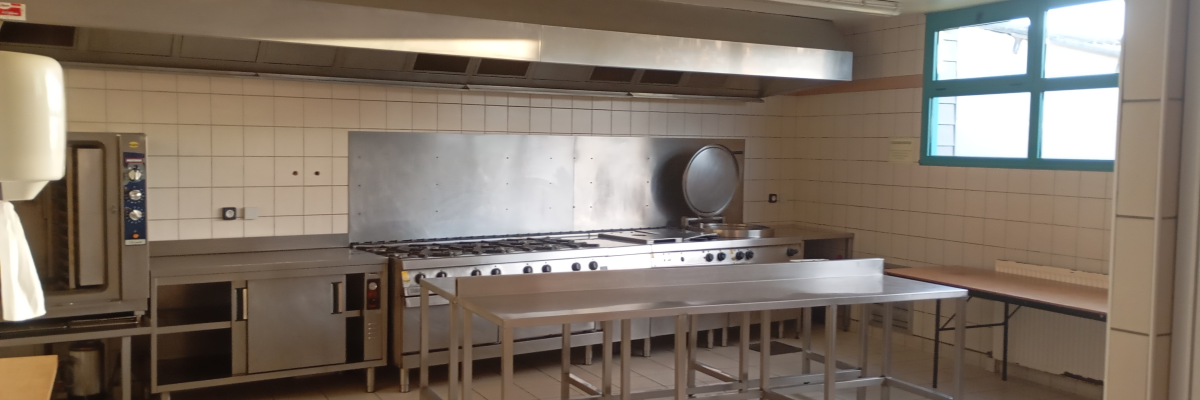 Cuisine professionnelle avec plans de travail, cuisinière, hottes, éviers, étagères et tables.