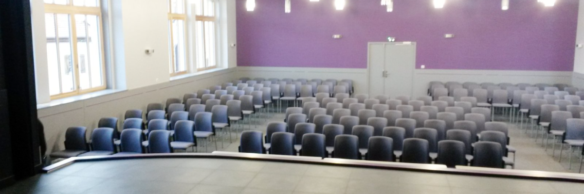 Salle avec rangées de chaises, scène, fenêtres, portes et éclairage suspendu.
