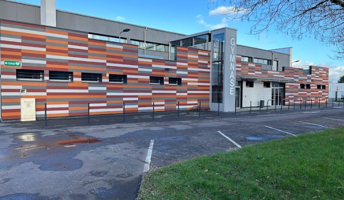 Bâtiment avec façade en briques colorées, parking et panneau "Gymnase".