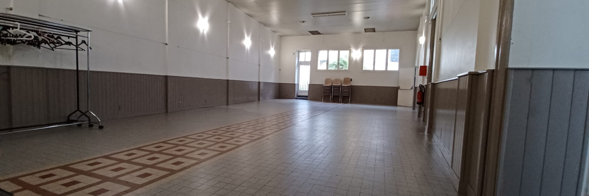 Grande salle vide avec portes, fenêtres, tapis de sol, rangement de chaises et climatiseurs muraux.