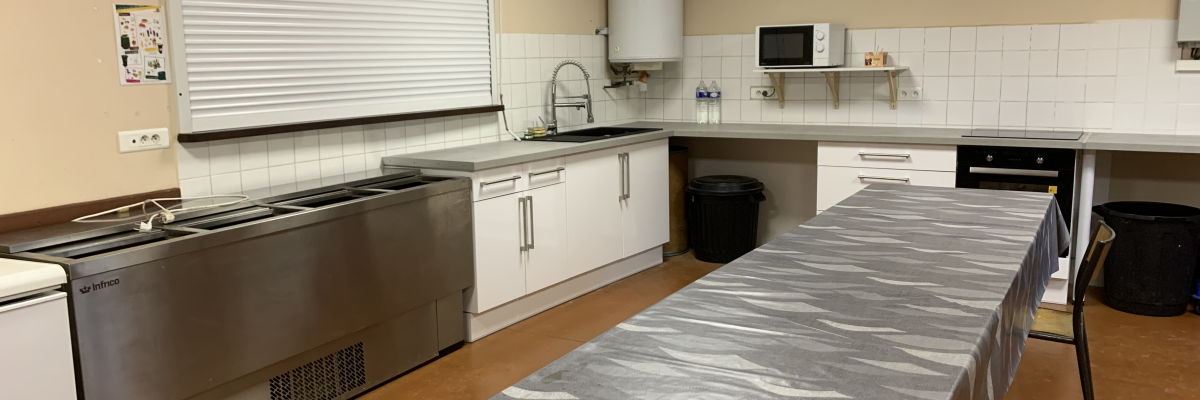 Cuisine avec plans de travail, évier, four, micro-ondes, réfrigérateur, table recouverte, poubelles et stores.