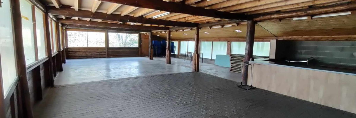 Salle spacieuse avec sol en brique, piliers en bois, grandes fenêtres, éclairage suspendu et mur en bois.