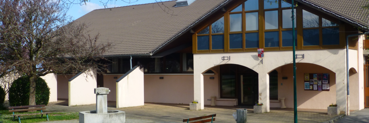 Bâtiment avec toit en pente, grandes fenêtres, porte d'entrée, abri extérieur, fontaine, banc et poubelle dans une zone herbeuse.