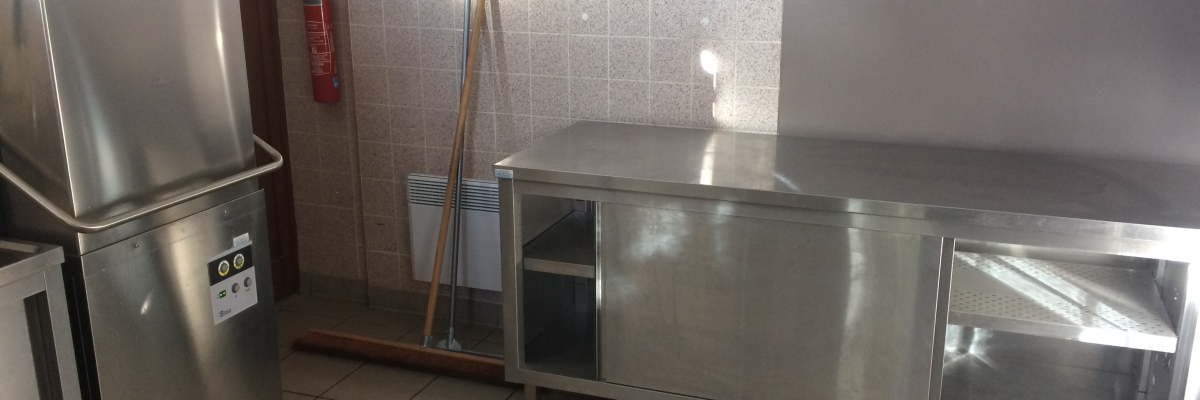 Cuisine avec lave-vaisselle, table inox, balai et extincteur.