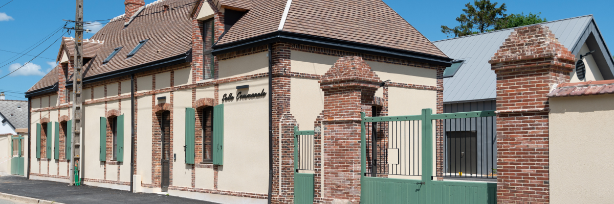 Maison en brique avec toit en tuiles, portes vertes, clôture et poteaux électriques.