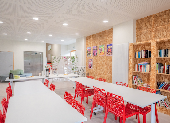 Salle avec tables, chaises rouges, étagères, livres, armoires et réfrigérateur.
