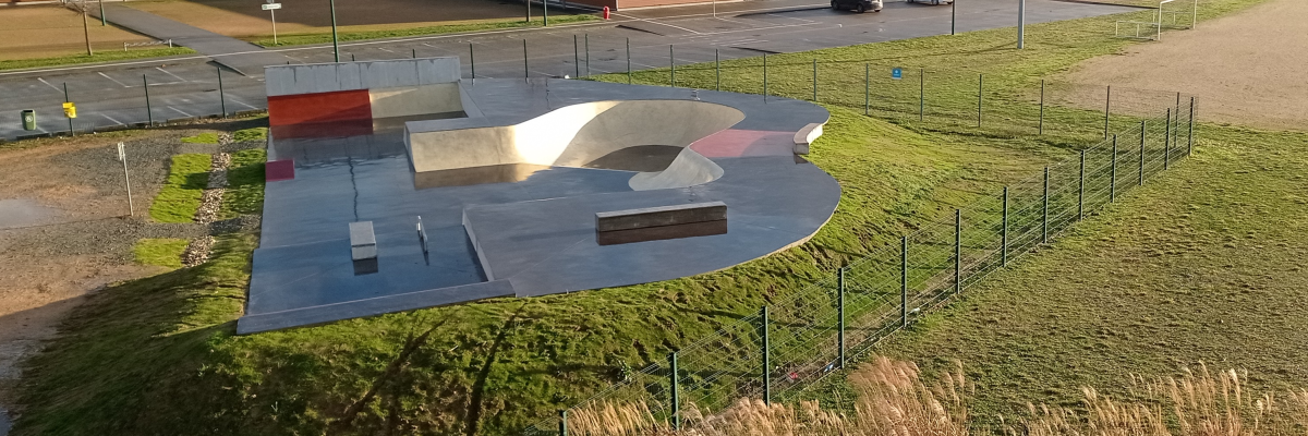 Skatepark avec rampes, clôture, parking et chemin.