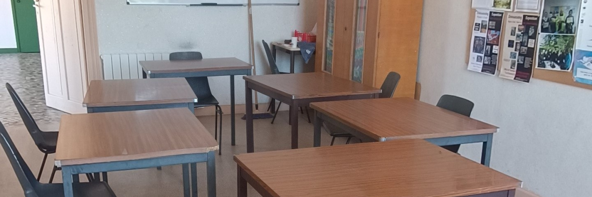 Salle de classe avec tables, chaises, tableau blanc, armoire et affiches.