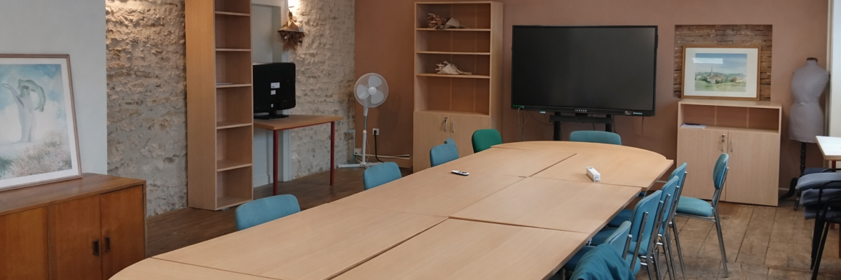 Salle de réunion avec table, chaises, écrans, ventilateur et étagères.