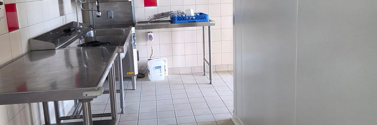 Cuisine avec plans de travail en acier inoxydable, éviers, étagères, balai et seau.