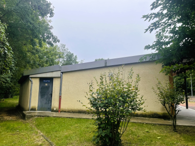 Bâtiment avec porte grise, escalier extérieur, buissons et arbres environnants.