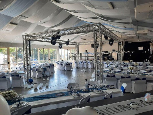 Salle de réception avec tables, chaises, structures métalliques, projecteurs, nappes et vaisselle.
