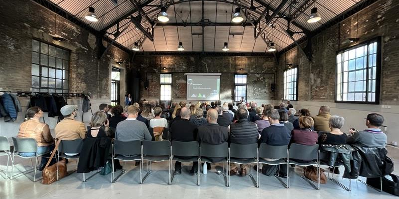 "Atelier avec chaises, projecteur, écran, public, grandes fenêtres et toit métallique."