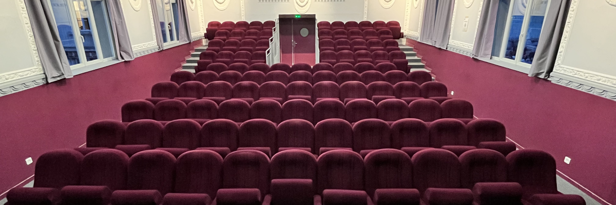 Salle avec rangées de sièges rouges, écran, projecteur et lumières au plafond.