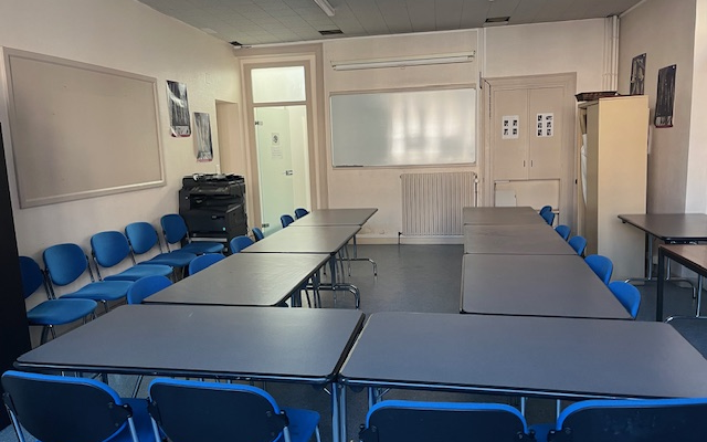 Salle de classe avec tables, chaises bleues, tableau blanc, porte, radiateur et tableau d'affichage.