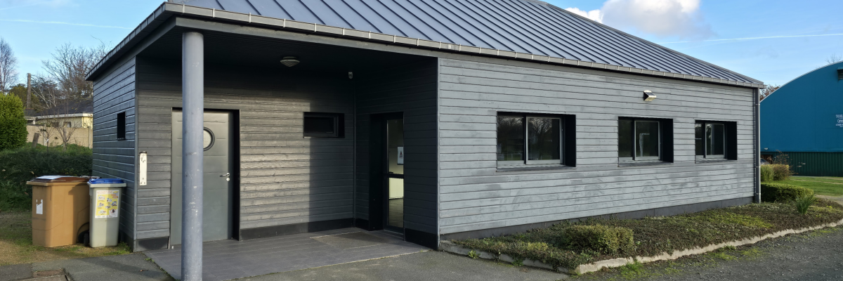 Bâtiment gris avec toit plat et colonnes, portes et fenêtres. Poubelles à côté. Ciel bleu avec nuages.