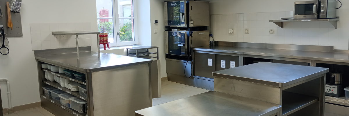 Cuisine professionnelle avec plans de travail, plaques de cuisson, four, hotte, évier, micro-ondes et étagères.