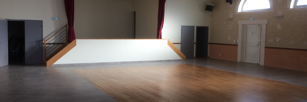 Salle avec scène, parquet, portes et fenêtres.