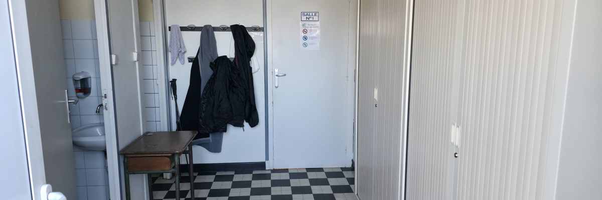 Couloir avec sol carrelé noir et blanc, porte, lavabo, urinoir, porte-manteau avec des vêtements et table.