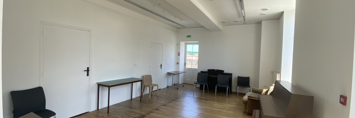 Salle avec tables, chaises, et porte. Sol en bois, murs blancs et grandes fenêtres.