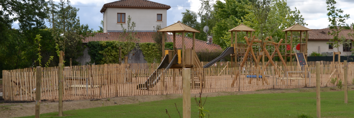 Aire de jeux avec toboggan, pont, et clôture en bois. Maison à l'arrière-plan.