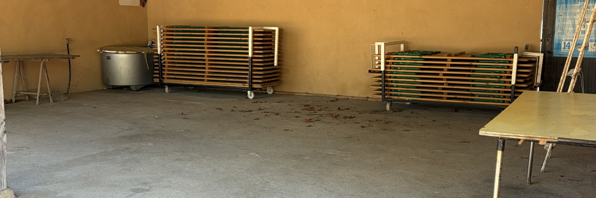 Salle avec tables, chaises, radiateurs et poubelle. Sol en béton, murs et plafond en bois.