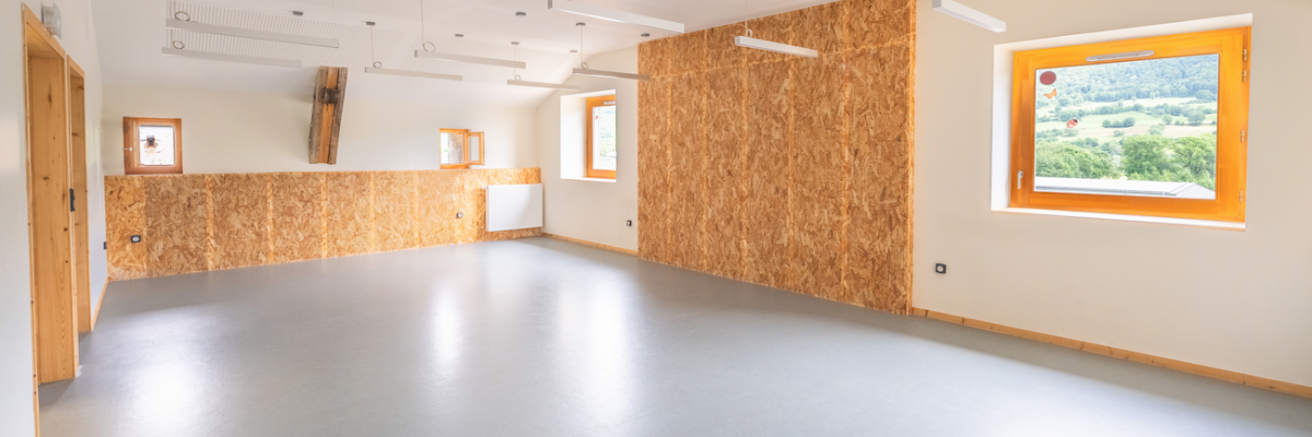 Salle vide avec murs en OSB, sol lisse, portes en bois, fenêtres et plafonniers.