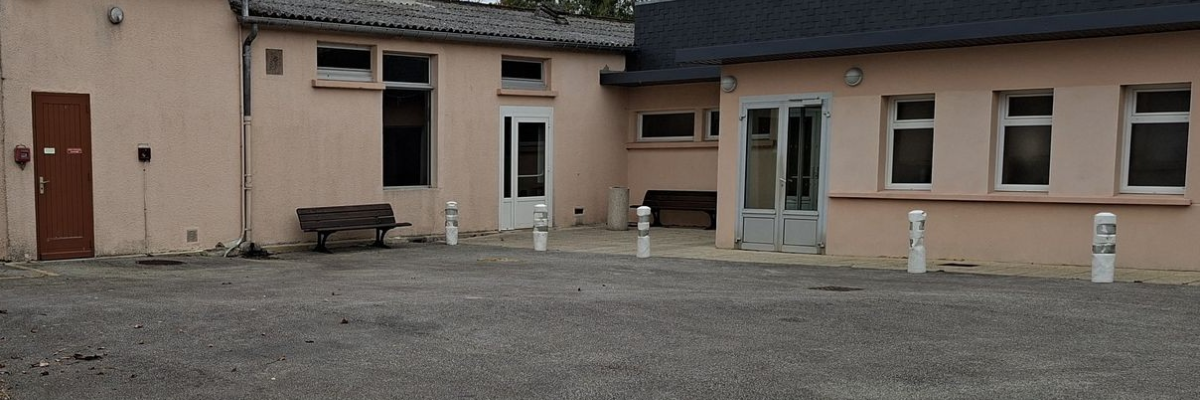 Bâtiment beige avec plusieurs fenêtres, porte en bois, banc et barrières.