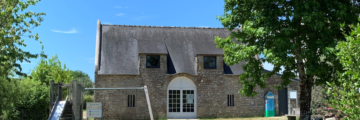 Maison ancienne avec toit en ardoise, aire de jeux extérieure, chemin de terre, poubelles, arbres et végétation.