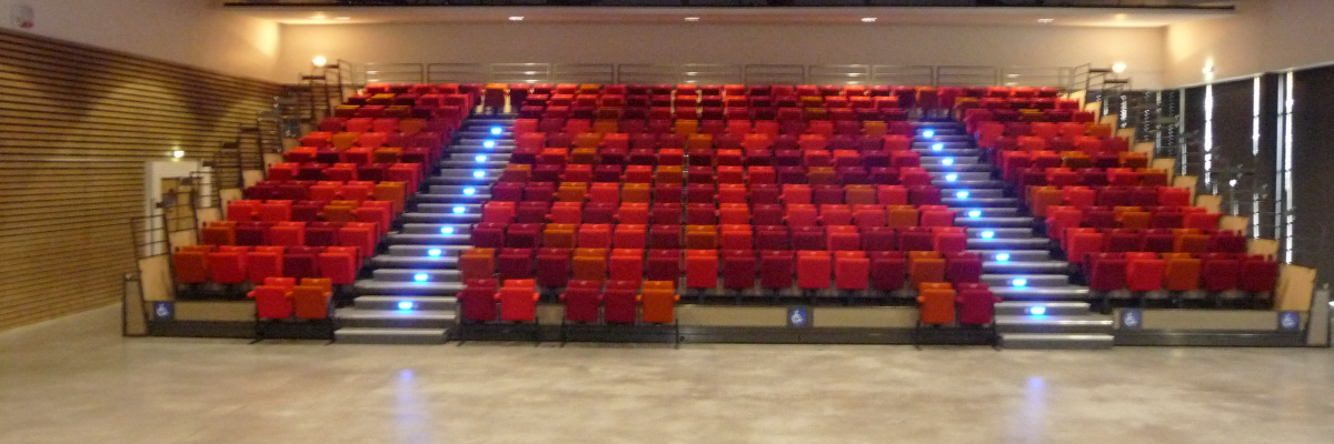 Salle avec rangées de sièges rouges, allée centrale, scène vide et éclairage au plafond.