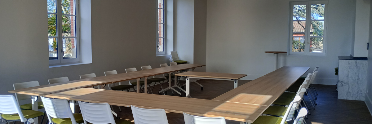 Salle avec longues tables, chaises, fenêtres, et lumière naturelle.