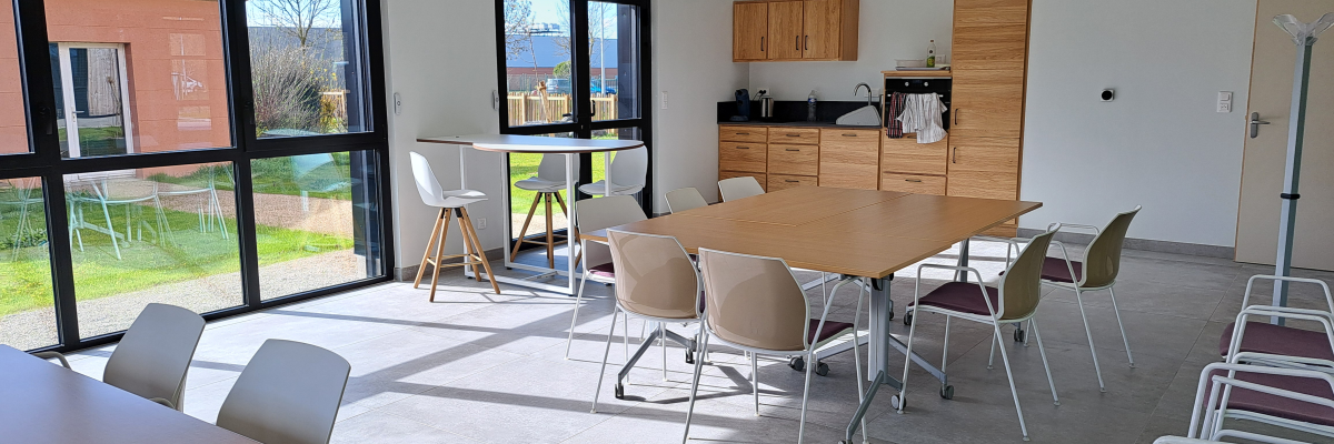 Salle avec tables, chaises, cuisine et grandes fenêtres.