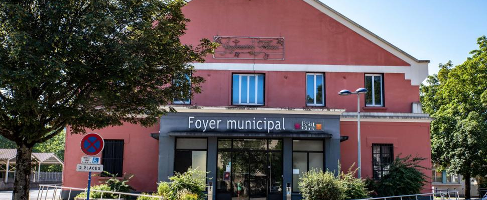 Foyer municipal, bâtiment rose à étage, entrée vitrée. Arbre à gauche, parking à droite.