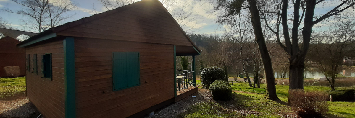 Cabane en bois avec fenêtres vertes, située dans un jardin avec arbres et gravier au sol.