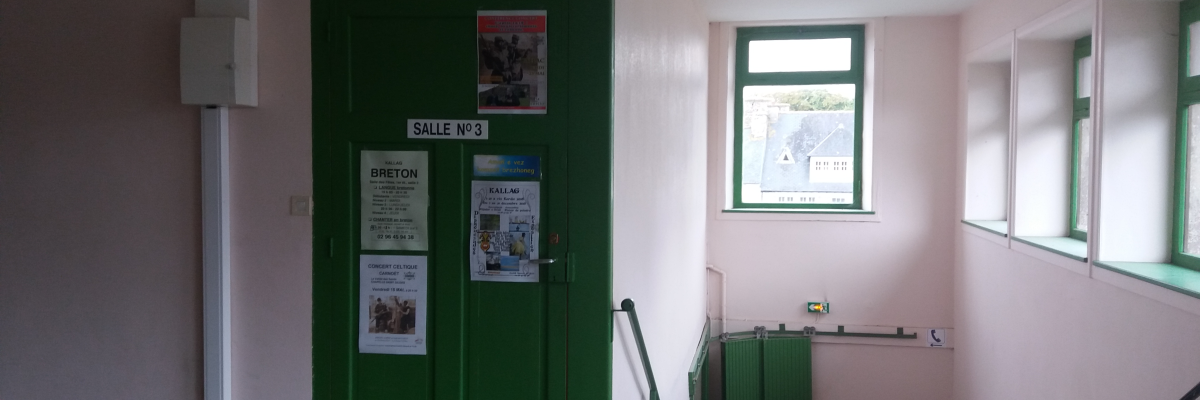 Pièce avec porte verte, fenêtre, affiches, radiateur et escalier.