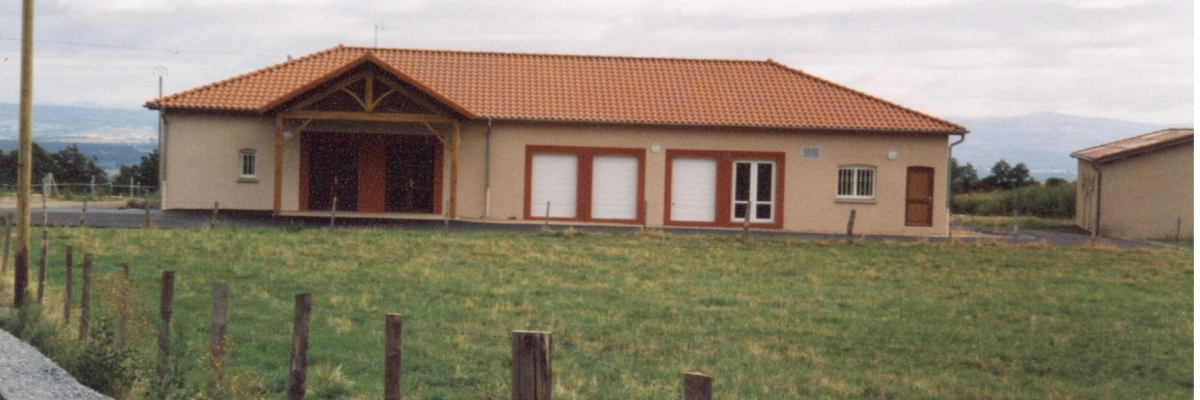 Bâtiment à toit orange, murs beiges, avec portes et fenêtres visibles, entouré d'une pelouse et clôturé. Route à gauche.