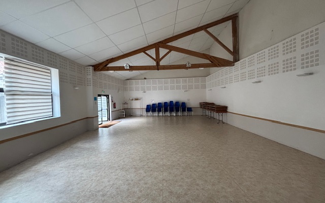 Salle vide avec chaises alignées, table, fenêtres et porte.