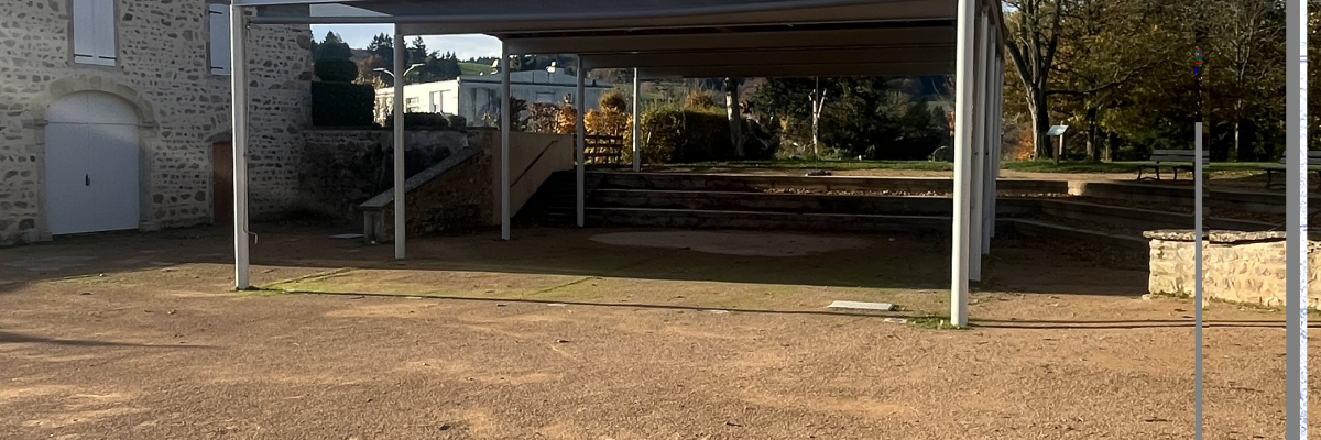 Structure en métal avec toit, terrain en terre battue, maison en pierre et arbres en arrière-plan.