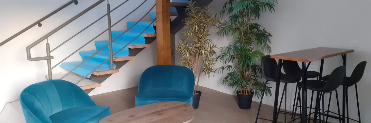 Espace avec escalier, fauteuils bleus, table basse, plantes, table haute et tabourets.