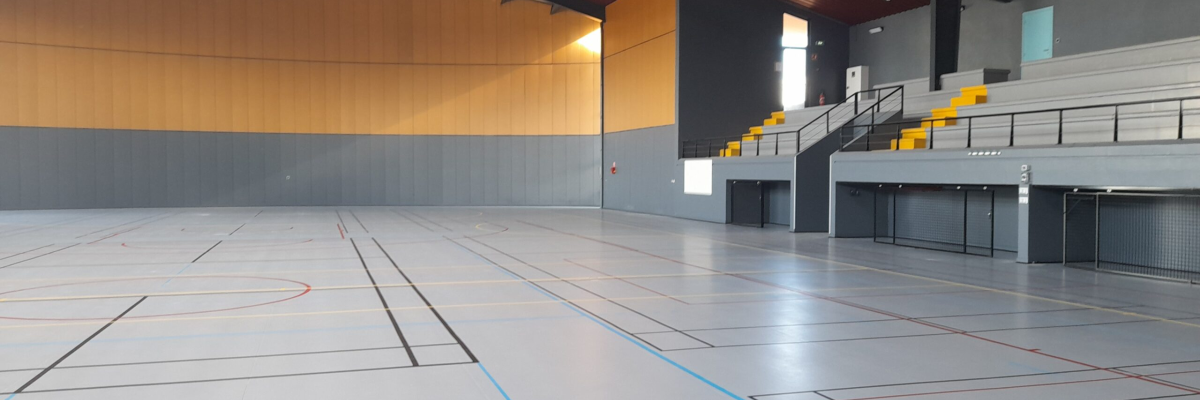 Salle polyvalente avec terrain de sport, gradins, murs métalliques et toit incliné.