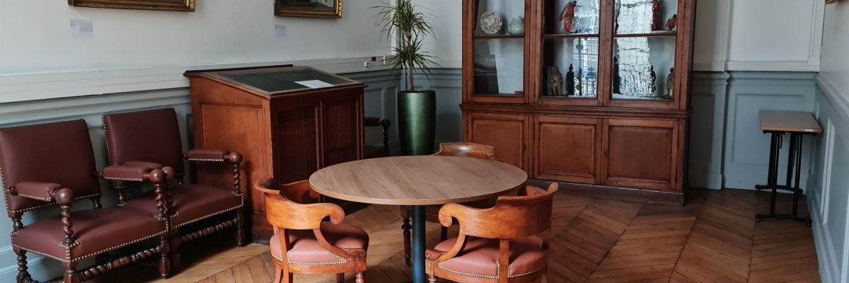 Salle avec table ronde, chaises, vitrine, banc et tableaux. Parquet au sol.