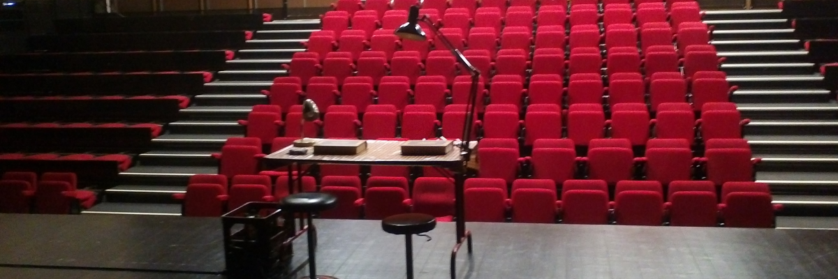 Salle avec scène, micro, tabouret, pupitre et rangées de sièges rouges.