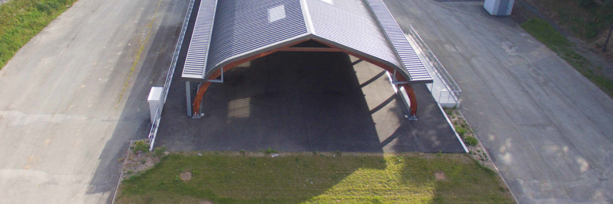 Hangar avec toit métallique, accès ouvert, aire de béton, graminées environnantes.