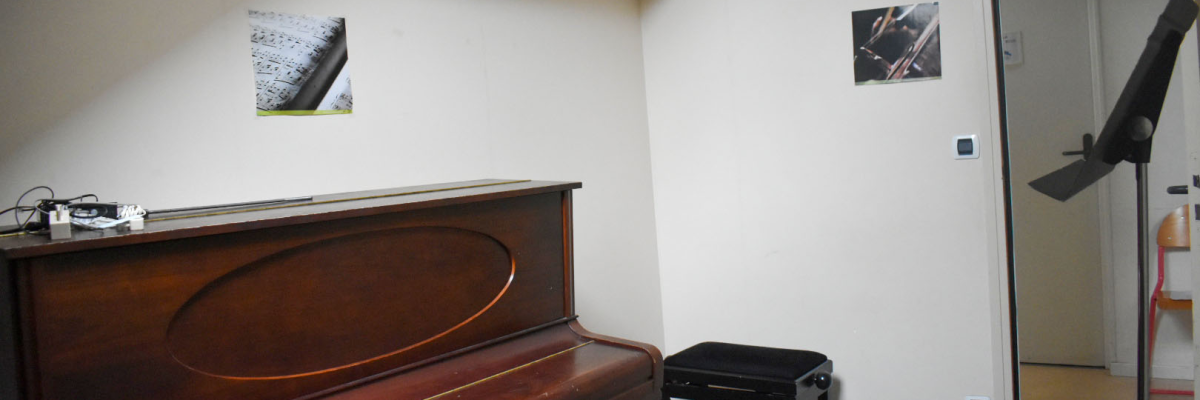 Pièce avec piano, tabouret et table basse. Deux cadres sur le mur. Porte ouverte avec comptoir visible.