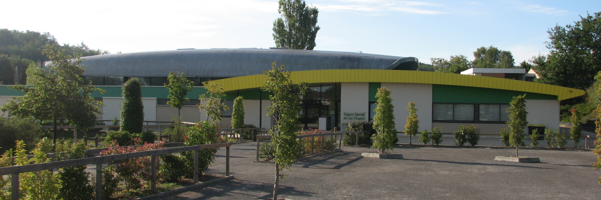 Bâtiment avec entrée circulaire, toiture incurvée, parking, arbres et fleurs.